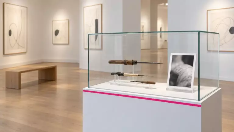 Heller Ausstellungsraum mit gerahmten Zeichnungen und einer Glasvitrine mit historischen Tätowierwerkzeugen und einer Schwarzweißfotografie